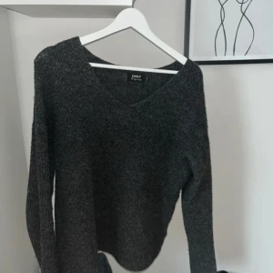 Mörkgrå stickad tröja från ONLY - Mysig mörkgrå stickad tröja från ONLY med v-ringad hals och lång ärm. Tröjan har en lite loose passform och är perfekt för kyliga dagar. Enkel att styla med jeans eller leggings för en avslappnad look.