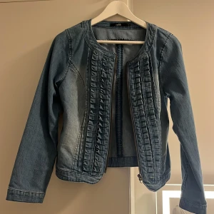 Jeansjacka med volangdetaljer Isolde - Snygg jeansjacka från Isolde i klassisk blå denim. Jackan har långa ärmar, rundad halsringning och dragkedja framtill. Framsidan är dekorerad med coola volangdetaljer som ger en unik touch. Perfekt för dig som vill sticka ut lite extra i vardagen.