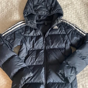 Mörkblå pufferjacka från Adidas S - Mörkblå pufferjacka från Adidas med huva och klassiska vita ränder på axlarna. Jackan är tillverkad i syntetmaterial som håller värmen. Perfekt för hösten och vintern!
