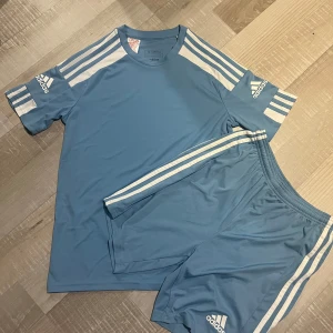 Blå Adidas dress med vita ränder - Snygga blå Adidas shorts med klassiska vita ränder längs sidorna och Adidas-logga framtill. Modellen har elastisk midja för skön passform och är gjorda i lätt polyester som andas. Perfekta för sport eller chill, matcha med t-shirt för en clean look. Man får med båda i priset! Ej hugget i sten