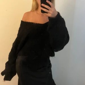 Svart offshoulder stickad tröja - Mysig svart stickad tröja med offshoulder-modell och bred ribbad kant vid halsen. Tröjan har en loose passform och långa ärmar med extra volym. Perfekt för dig som gillar en avslappnad men ändå trendig look.