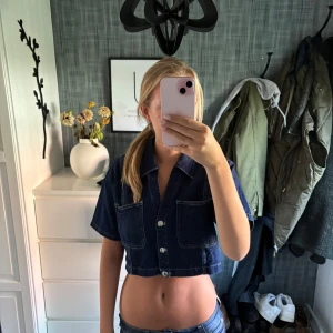 Mörkblå croppad jeansskjorta - Säljer en mörkblå croppad skjorta i jeansmaterial med korta ärmar och vita knappar framtill. Skjortan har två stora bröstfickor och en klassisk krage. Perfekt för dig som gillar en chill och trendig stil.