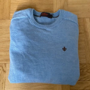 Ljusblå merinoullströja från Morris - Säljer en stilren ljusblå tröja från Morris i 100% merinoull. Tröjan har rund halsringning, ribbade muddar och en liten broderad logga på bröstet. Perfekt för dig som gillar klassisk och clean stil.