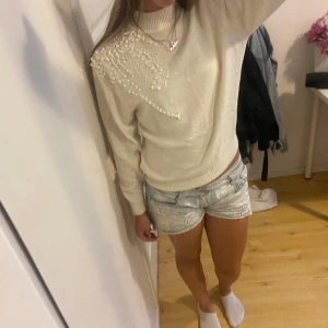 Vit stickad tröja med broderi - Superfin vit stickad tröja med hög hals och unikt broderat blommönster på ena axeln. Tröjan har långa ärmar och ribbade muddar, perfekt för att ge en mysig och stilren look. Passar dig som gillar detaljer och vill sticka ut lite extra.
