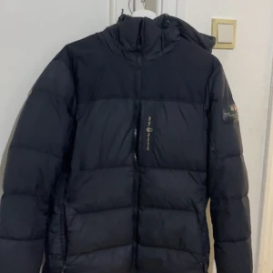 Svart dunjacka från Geographical Norway - Säljer en svart dunjacka från Geographical Norway med huva och dragkedja. Jackan har långa ärmar och är perfekt för kalla dagar. Den har en stilren design med märkets logga på bröstet.salingrace jacka 