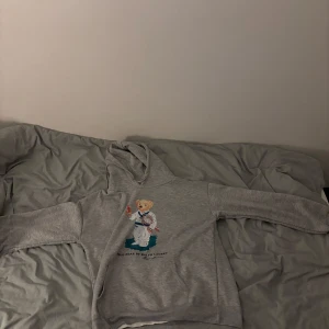 Grå hoodie från Ralph Lauren med björnmotiv - Säljer en grå hoodie från Ralph Lauren med ett charmigt björnmotiv på framsidan. Hoodien har en klassisk design med långa ärmar och en bekväm passform. Perfekt för en avslappnad stil.