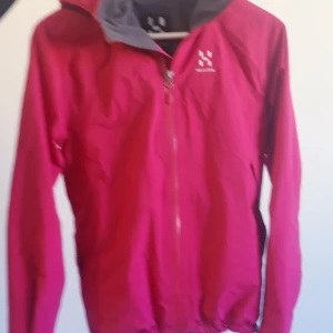 Rosa vindjacka från Haglöfs - Säljer en rosa vindjacka från Haglöfs med dragkedja och huva. Jackan har en grå insida och är perfekt för blåsiga dagar. Den är lätt och smidig att bära med sig.