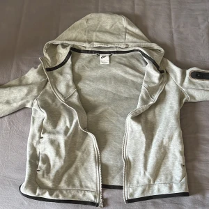 Grå huvtröja från Nike - Säljer en grå huvtröja från Nike med dragkedja och huva. Tröjan har svarta detaljer och en ficka på ärmen. Perfekt för en sportig look eller avslappnade dagar. Storlek 158-170.