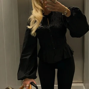 Svart blus med volangdetaljer - Snygg svart blus med smockdetaljer och volang i nederkant. Blusen har långa ärmar och en knäppning framtill. Perfekt för en stilren look.