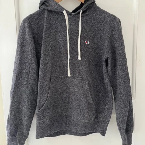 Grå hoodie från Champion - Säljer en klassisk grå hoodie från Champion med vit snörning och det ikoniska Champion-logotypen på bröstet. Perfekt för en avslappnad stil. Den har en stor ficka framtill och är tillverkad i mjukt material för extra komfort. Står att det är storlek M men den sitter som S