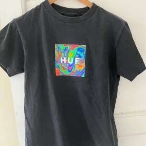 Svart t-shirt från HUF - Säljer en skitcool svart t-shirt från HUF med ett färgglatt, psykedeliskt mönster på bröstet. T-shirten är kortärmad och har en klassisk rund halsringning. Perfekt för en avslappnad stil.