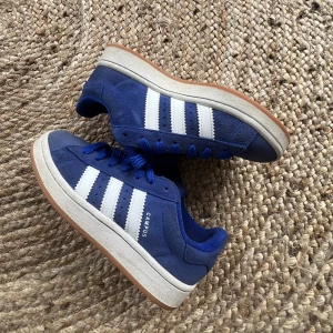 Blå Adidas Campus sneakers - Snygga blå Adidas Campus sneakers med vita ränder och gummisula. Skorna har en klassisk design med snörning och är tillverkade i mocka för en stilren look. De är väl använda men i ganska bra skick. Storlek 38💘 PRIS KAN DISKUTERAS!!