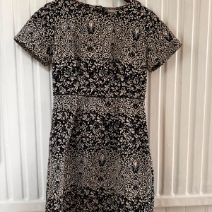 Mönstrad klänning från H&M - Säljer en elegant mönstrad klänning från H&M i svart och beige jacquardvävd med blommönster. Klänningen har korta ärmar och en rund halsringning. Perfekt för en stilfull look!