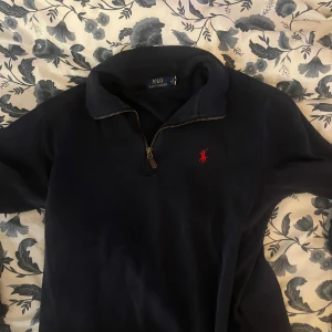 Mörkblå tröja från Polo Ralph Lauren - Säljer en stilren mörkblå tröja från Polo Ralph Lauren med dragkedja och klassisk krage. Den har en broderad röd logotyp på bröstet. Perfekt för en avslappnad och snygg look.