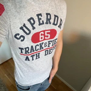 Grå t-shirt från Superdry - Säljer en grå t-shirt från Superdry med tryck i rött och svart. T-shirten har korta ärmar och texten 'Track & Field' på framsidan. Perfekt för sommaren.