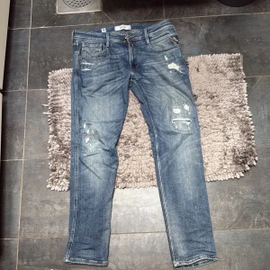 Replay jeans anbass - Säljer ett par blå jeansbyxor från Levis med slitna detaljer och en klassisk femficksdesign. Byxorna har en normal passform och är perfekta för en avslappnad stil. De har en dragkedja och knapp framtill.