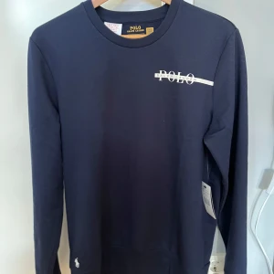 Mörkblå tröja från Ralph Lauren - Säljer en stilren mörkblå tröja från Ralph Lauren med vit POLO-logga på bröstet. Tröjan har långa ärmar och en klassisk rund halsringning. Perfekt för en avslappnad och snygg look.