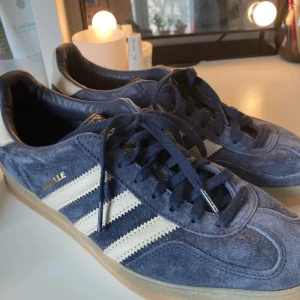 Adidas Gazelle i blå mocka - Snygga Adidas Gazelle sneakers i blå mocka med vita ränder och snörning. Perfekta för en avslappnad stil med en klassisk touch. De har en gummisula som ger bra grepp och komfort.
