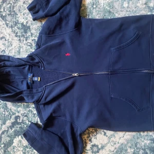 Mörkblå Polo Ralph Lauren zip tröja  - Säljer en snygg mörkblå huvtröja från Polo Ralph Lauren med dragkedja och broderad röd logotyp på bröstet. Tröjan har långa ärmar och en praktisk huva. Perfekt för en avslappnad stil.