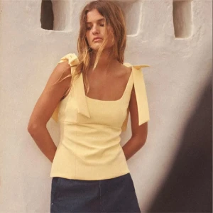 Gul  topp med rosetter - Snygg gul topp med stora rosetter på axlarna. Perfekt för sommardagar och ger en fräsch look. Toppen har en enkel och stilren design som passar till många olika outfits.