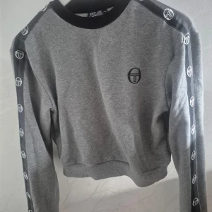 Grå sweatshirt från Sergio Tacchini - Säljer en grå sweatshirt från Sergio Tacchini med svarta detaljer och logotyp längs ärmarna. Tröjan har en rund halsringning och ribbade muddar. Perfekt för en sportig look!