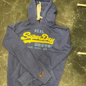Superdry Hoodie - Säljer en blå hoodie från Superdry med gul och blå text på framsidan. Tröjan har en justerbar huva med vita snören och en stor ficka framtill. Perfekt för en avslappnad stil.