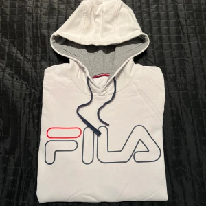 Hoodie från FILA - Säljer en vit hoodie från FILA med stor logga i blått och rött på framsidan. Hoodien har en justerbar huva med dragsko och en praktisk magficka. Perfekt för en avslappnad stil.