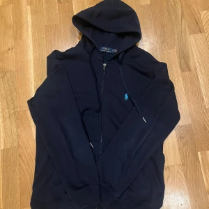 Mörkblå hoodie från Polo Ralph Lauren - Säljer en snygg mörkblå hoodie från Polo Ralph Lauren med dragkedja och huva. Den har en liten broderad logga på bröstet och är perfekt för en avslappnad stil. Passar bra till både jeans och joggers.