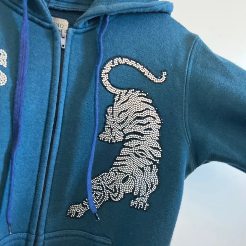 Snygg blå hoodie från Vansydical med en cool tigerdesign i strass på framsidan. Tröjan har dragkedja och justerbara snören i huvan. Perfekt för en avslappnad stil.. Neuletakit & Villapaidat.