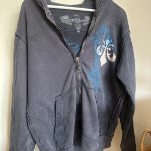 Grå/marin blå hoodie med dödskallemotiv från Affliction - Säljer en grå hoodie från Affliction med ett stort dödskallemotiv och blåa detaljer. Tröjan har dragkedja och en avslappnad passform. Är inte säker vilken storlek den är men gissar på L💕 Kan användas både av män och kvinnor. Tröjan har tre störra skador dem finns på de tre sista bilderna, men skadorna märks inte eftersom de passar till stillen🤗