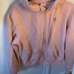 Rosa hoodie med hjärtdetalj - Mysig rosa hoodie från H&M med en liten hjärtdetalj på bröstet. Tröjan har en klassisk känguruficka och en bekväm huva. Perfekt för en avslappnad stil. Storlek S . Säljer pga den ligger bara i garderoben och tar plats ,köpt för länge sen för cirka 250 kronor.