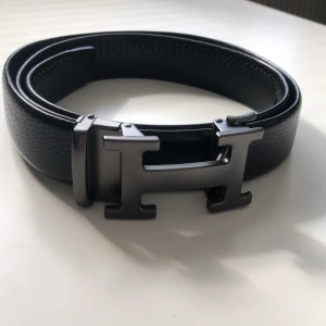 Svart bälte med H-spänne - Snyggt svart bälte med ett stilrent H-format spänne i metall. Perfekt för att ge en elegant touch till din outfit. Tillverkat i skinn för en lyxig känsla.