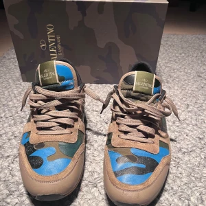 Valentino Rockrunner sneakers - Snygga Valentino Rockrunner sneakers med ett unikt kamouflagemönster i blått, grönt och beige. Skorna har snörning och en cool design med nitar på hälen. Perfekta för dig som vill sticka ut med stil.