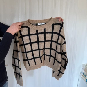 Beige och svart rutig stickad tröja från NAKD - Säljer en snygg beige stickad tröja från NAKD med ett svart rutmönster. Tröjan har långa ärmar och en rund halsringning. Perfekt för kyligare dagar!