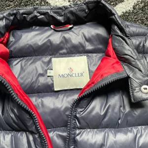 Mörkblå pufferjacka från Moncler - Säljer en snygg mörkblå pufferjacka från Moncler med röd insida. Jackan har en dragkedja framtill och Moncler-logga på ärmen. Perfekt för vår dagar eller kalla sommardagar den har lite defekter inifrån men inget som man märker.