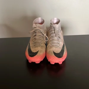 Nike Mercurial fotbollsskor - Snygga Nike Mercurial fotbollsskor i beige och rosa med svarta detaljer. Skorna har en hög ankelkrage och snörning för bra passform. Perfekta för fotbollsplanen med sina greppvänliga dobbar.