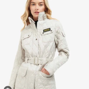 Oanvänd quiltad jacka från Barbour International XS - Snygg beige quiltad jacka från Barbour med bälte i midjan och flera fickor. Jackan har en hög krage med spänne och knappar framtill. Perfekt för kyligare dagar.  Helt ny och oanvänd med tag kvar. 