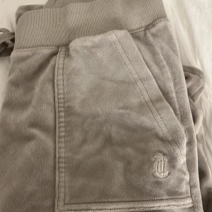 Juicy byxor - Säljer ett par beige mjukisbyxor med ribbad midja och snörning. Byxorna har en broderad logga på framsidan och bakfickor med knappar. Bra skick säljer pga för små