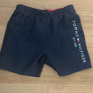 Svarta shorts från Tommy Hilfiger - Snygga mörkblå badshorts från Tommy Hilfiger med logotyptryck på sidan. De har en elastisk midja med snörning och är perfekta för sommarens äventyr. Insidan har ett rött foder och ett nät för extra komfort. Storlek 14-16