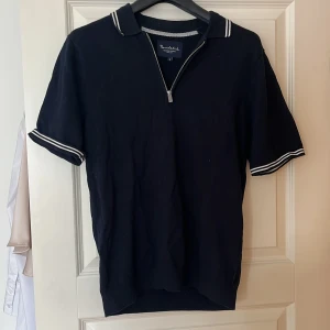 Svart pikétröja från Bondelid - Snygg mörkblå pikétröja från Bondelid med dragkedja och vita ränder på krage och ärmslut. Perfekt för en stilren look.