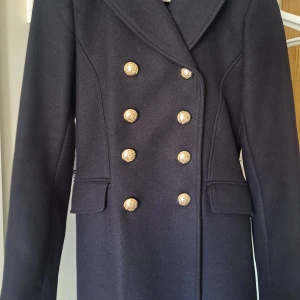 Mörkblå skepparjacka med guldknappar - Elegant mörkblå skeppar jacka/ kappa med dubbla rader av guldknappar framtill och på ärmarna. Kappan har en klassisk design med långa ärmar och fickor med lock. Perfekt för en stilren look. Liten i storlek.