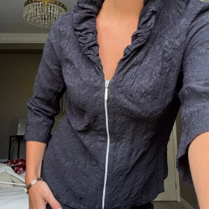 Spets kofta - Snygg svart jacka från Stine Goya med volangkrage och dragkedja framtill. Jackan har ett diskret spetsmönster och trekvartsärmar, vilket ger en elegant touch. Perfekt för att lyfta din outfit eller bära för sig!