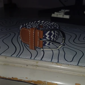 Randigt bälte med brunt läder - Snyggt randigt bälte i blått och vitt med detaljer i brunt läder. Perfekt för att ge en stilren touch till din outfit. Bältet har ett klassiskt spänne i metall. De är helt oanvänd och orkar inte skicka tillbaka den ee är typ 70 cm långt men är utsträckbar skriv om du har några frågor eller funderingar! 