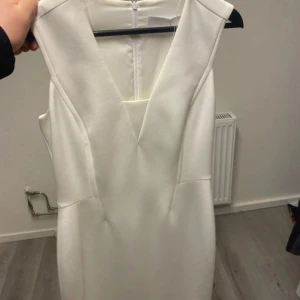 Vit klänning från HUGO BOSS - Elegant vit klänning från BOSS med v-ringad design och dragkedja i ryggen. Perfekt för en stilren look. Klänningen har en figurnära passform och är ärmlös, vilket ger en modern och sofistikerad känsla.