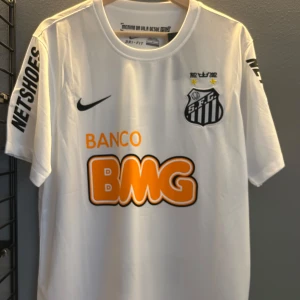 Vit fotbollströja från Nike - Säljer en vit fotbollströja från Nike med korta ärmar. Tröjan har tryck i svart och orange, inklusive ett klubbmärke och sponsorloggor. Perfekt för fotbollsfans som vill visa sitt stöd. Storlek L och en replika helt ny med tagg