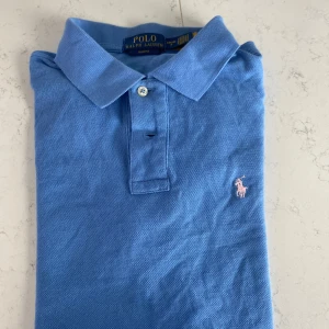 Blå pikétröja från Ralph Lauren - Säljer en klassisk blå pikétröja från Ralph Lauren med den ikoniska loggan broderad i rosa på bröstet. Tröjan har en krage och knappar framtill. Perfekt för en stilren look.