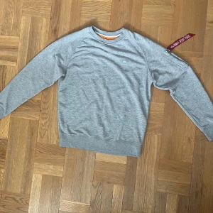 Grå tröja från Alpha Industries - Säljer en stilren grå tröja från Alpha Industries med en liten logga på bröstet. Tröjan har långa ärmar och en dragkedja på armen. Insidan av kragen är orange vilket ger en cool kontrast. Perfekt för en casual look! 9/10 inga defekter! Knappt använd. Info för mer bilder/detaljerade bilder