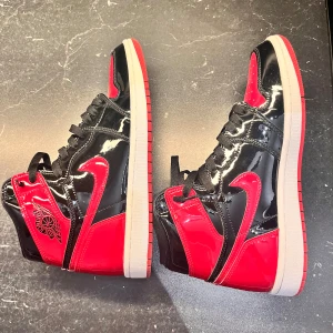 Jordan 1 high patent bred - Snygga Nike Air Jordans i en iögonfallande kombination av svart och röd lack. Skorna har klassisk snörning och en rund tå. Perfekta för den som vill sticka ut med stil. Storlek 44