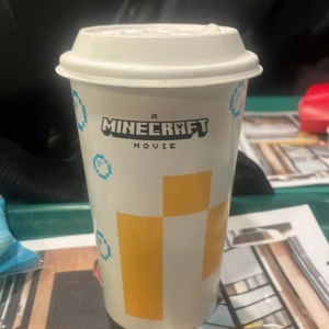 Minecraft Movie Pappersmugg - En pappersmugg från McDonald's med ett Minecraft Movie-tema. Muggen har ett tryck med blå pixlade bubblor och en stor gul rektangel. Locket är vitt med olika symboler för dryckesval. Bra skick 