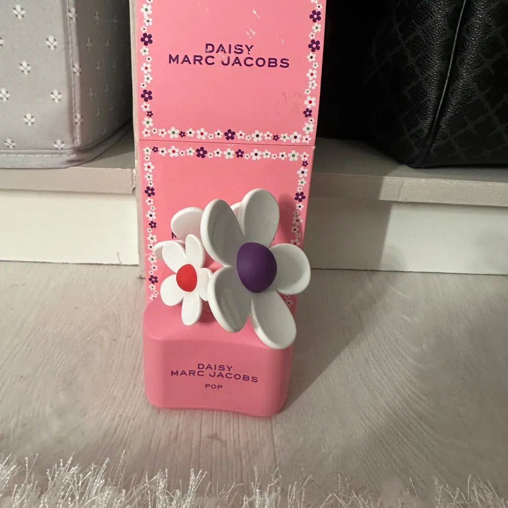 Säljer en Daisy Marc Jacobs Pop parfym i en söt rosa flaska med blommiga detaljer. Flaskan har en unik design med vita blommor och lila samt röda detaljer på korken. Perfekt för den som älskar en lekfull och fräsch doft.. Perfume.
